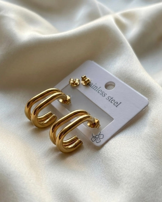 Gold hoop earrings on a white card with ' stainless steel' text, placed on a beige fabric background.