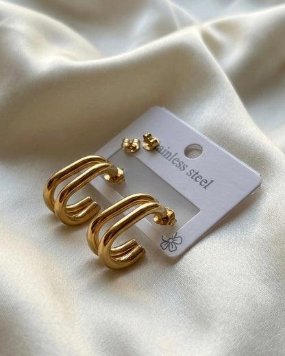 Gold hoop earrings on a white card with ' stainless steel' text, placed on a beige fabric background.